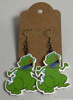 Pair of green dinosaur dangle earrings mounted on a brown cardboard display card, new condition.