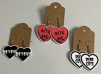 Photo of three pairs of Halloween-themed heart-shaped dangle earrings. The earrings are black with 'PSYCHO', red with 'BITE ME', and white scalloped with 'DEAD CUTE' sayings. Each pair is attached to a brown earring card.