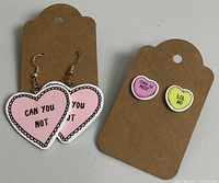 Two pairs of heart shaped earrings displayed on brown earring cards: one pair of pink dangle hearts with 'CAN YOU NOT' text, and one pair of small stud hearts with 'CAN U NOT' and 'LOL NO' text.