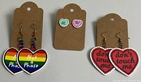 Three pairs of heart-shaped earrings displayed on brown earring cards: rainbow multicolor dangle earrings, small heart stud earrings with text, and red dangle earrings with text.