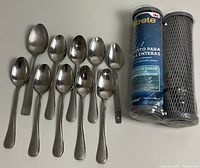 Two Filtrete water filters (one in packaging, one out) plus ten stainless steel spoons (five tablespoons and five teaspoons) laid out on white surface.