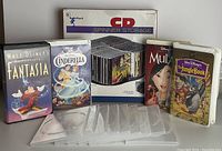 Photo shows four Walt Disney VHS tapes - Fantasia, Cinderella, Mulan, Jungle Book - beside a Radio Shack branded CD spinner storage box containing multiple CDs, and several empty clear plastic disc cases.