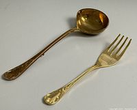 Photo of a large gold tone serving fork and ladle placed side by side on a white background showing detailed handle design.