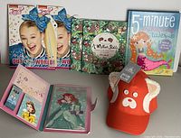 Photo showing 2 Jojo Siwa diaries, 2 A Million Sloths notebooks, 1 Enchanting Tales 5-minute storybook, 1 Disney Princess magnetic book, and 1 orange cap with animal ears