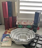 Wide shot showing collection of holiday napkins, six tall holiday tin containers in red, blue, and gold, nestable metal flower-shaped baking pans, metal brownie pan with nine square sections, three rectangular holiday-themed treat containers, green holiday travel cup, cookie cutters, wooden utensils, and measuring spoon.
