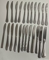 Photo of 22 kitchen knives and spreader knives laid out in three rows on a white surface. The knives have stainless steel blades and handles with different designs. Some have smooth rounded handles, some have ornate patterned handles. All knives show signs of use.