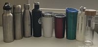 Photo of 8 drinkware items arranged in a row on a white surface including insulated bottles, tumblers, a pitcher and a glass water bottle