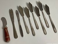 Photo showing 8 assorted knives and utensils including a red wooden handle knife and various stainless steel spreaders and knives with different blade shapes and handle designs.