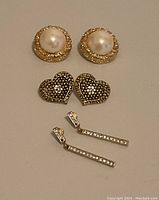 Three pairs of vintage earrings arranged on a plain background: faux pearl button earrings, heart-shaped rhinestone earrings, and rectangular rhinestone dangle earrings.