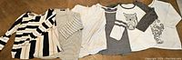 Six women's plus size shirts in grey, white, and black displayed on a wooden surface. Designs include stripes, solid colors, and graphic prints.
