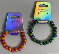 Two handmade wooden beaded bracelets on holographic cards, one with pink, orange, and yellow beads, the other with green and purple beads.