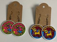 Photo of two pairs of zodiac-themed dangle earrings, one Taurus and one Sagittarius, displayed on plain brown cardboard earring holders against a white background.