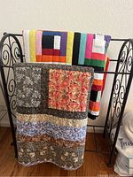 Three handmade quilts displayed on a black metal quilt rack. The front quilt shows a floral and leaf pattern with detailed patchwork.