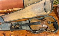Antique violin seated inside a worn gray-lined hard violin case, showing the full instrument and case interior.