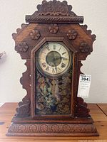 Front view of antique gingerbread mantle clock showing carved wooden frame, Roman numeral clock face, and floral decorative glass door.