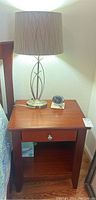 Full view of a wood bedside table with metal lamp and cream shade on top, clock beside lamp, and drawer closed.