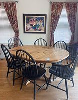 Oval wooden dining table with six black Windsor style chairs arranged around it in a room with wood floor and patterned curtains.