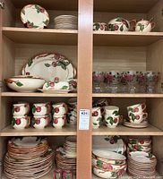 Wooden cabinet shelves filled with assorted Franciscan Apple dinnerware including plates, bowls, cups, saucers, serving platters, and glasses.