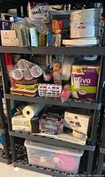 Full shelving unit with multiple shelves containing various disposable paper products and dishware including plates, cutlery, cups, napkins, and straws.