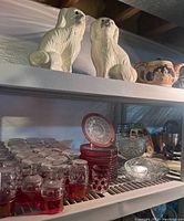 Photo showing white ceramic dog figurines, ceramic teapot, cranberry thumbprint glassware including small glasses and plates stacked on white shelving.