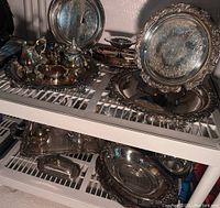 Overview of multiple Wallace silverplate trays and tea set pieces placed on shelving.