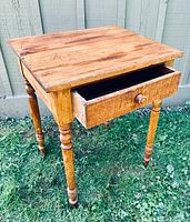 View of antique wooden side table showing the bird's eye maple top, turned legs, and open drawer.