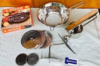 Full set of kitchen tools displayed: cast iron dough press (boxed and unpacked), stainless steel food mill with handle and three grinding discs, all laid out on a white cloth surface.