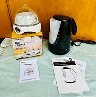 White egg cooker with transparent lid placed on top of its orange and white box, instruction manual laid out on table beside it, alongside a black electric kettle with manual.