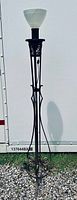 Full view of wrought iron torchiere floor lamp standing outside on gravel against a white wall, showing its black iron frame and milk glass shade.