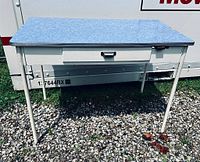 Full view of Mid-century modern blue formica table desk with white metal legs and one front drawer, standing outdoors on gravel.