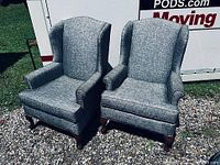 Front view of both armchairs side by side on gravel with moving pod in background. Shows overall shape, upholstery and wood legs.