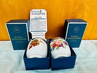 Two porcelain Royal Worcester egg coddlers seated in original dark blue boxes, one decorated with red berries, the other with floral and pink rose design. Instruction leaflet displayed behind.