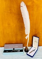 Photo shows a white feather quill affixed upright in a small vintage glass inkwell with a cork, alongside a Pierre Belvédère ballpoint pen in a brown box and a Fisher Space pen in a blue box.