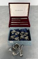 Photo showing original red velvet-lined case with 4 American Godinger silver spreaders and a box with 4 Seagull pewter bow-shaped napkin rings with blue tissue lining below.