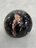 Top and angled view of one black glass paperweight with copper-colored inclusions inside. Reflective surface and smooth finish evident.