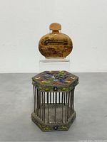 Shows the full set: the snuff bottle on a clear stand behind a small hexagonal, enamel-decorated Asian birdcage with metal bars