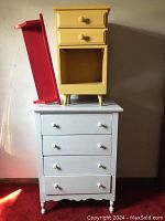 Full view showing white dresser with four drawers, yellow nightstand placed on top, and red wall shelf leaning against nightstand
