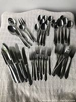 Overview of multiple forks, knives, and spoons in two patterns spread on white towel, showing polished stainless steel flatware assortment.