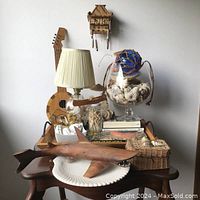 Entire lot displayed on wooden side table, including carved wood shark on oyster shell plate, lamp, glass snifter with shells, books, wooden instrument, and woven boxes