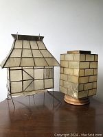 Two vintage capiz shell lamps, one hut-shaped with bent metal, the other square on wooden base, placed on a wooden table.