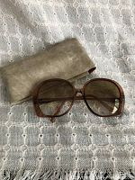 Photo showing oversized Bella Paris sunglasses frames and their soft beige case on a textured fabric background.