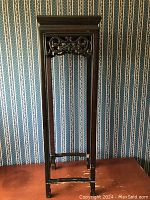 Vintage Chinese rosewood plant stand tall table, square top with carved scrollwork gallery beneath, dark stained wood, standing against wallpaper.