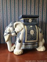 Side view of the vintage white ceramic elephant garden stool with blue decorative detailing showing the raised design on the blanket and saddle, and trunk down position.