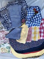 Photo shows various folded placemats and napkins including blue and white gingham, yellow and red checkered, and blue geometric patterns along with a piece with scalloped edges and embroidery.