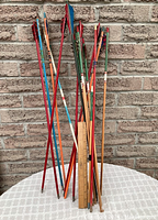 Photo of assorted vintage arrows with multicolor fletching standing upright on a white surface against a brick wall, showing a tape measure for scale.