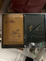 Two A.A. Milne books in plastic drawer: Now We Are Six (yellow cover) and black hardcover with gold illustration