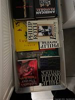 Clear top view of plastic bin filled with Philip Roth books including 'Harmony', 'Sabbath's Theater', 'Operation Shylock' and others