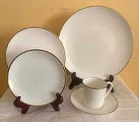 White ceramic dinnerware set with three plate sizes and matching tea cup with saucer, all with gold rim detail.