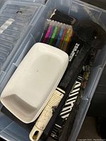 Plastic container holding various art supplies including charcoal sticks, colored markers, a paintbrush, and a white tray.