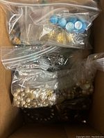 Box containing multiple clear plastic bags with various jewelry pieces including faux pearls, metal chains, and beads.